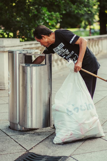 A middle-aged man with dark hair, wearing a black T-shirt with white text and dark trousers, is bent over on a paved sidewalk, placing a large white plastic bag filled with waste into an open-top, sta