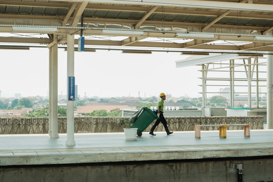 A middle-aged man with dark hair, wearing a black T-shirt with white text and dark trousers, is bent over on a paved sidewalk, placing a large white plastic bag filled with waste into an open-top, sta