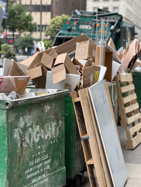 A green outdoor clothing donation and recycling box positioned against a backdrop of dense foliage and a wooden fence. The box features Chinese characters and a phone number on its front, with designa